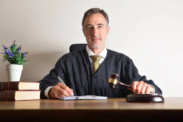Judge dressed in the toga and with the gavel in his hand signing documents in his office on wooden table and isolated light background. Front view