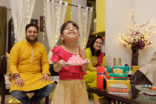 Cute little Kid, dressed up in ethnic wear holding plate of sweets with parents seating in the back expressing emotion looking at the candle celebrating diwali Hindu festival Laxmi poojan