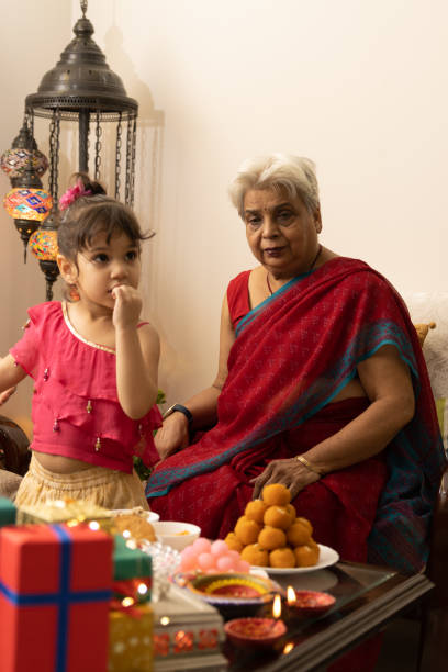 A cute little girl kid dressed up in ethnic standing next to grandmother in saree celebrating diwali Hindu festival with sweets and gifts stock photo