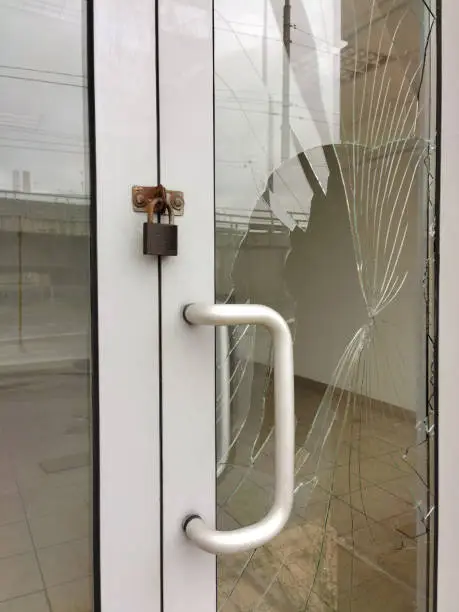 Photo of The glass of a store door broken by thieves or vandals with a padlock hanging from it