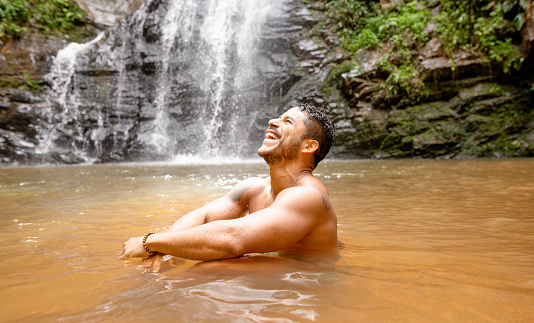 Fit young man laughing while relaxing in the pool of a waterfall in a tropical forest