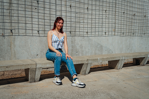 Beautiful latin woman with red hair sitting on a cement bench wearing sunglasses and wearing a jean and a sleeveless top on a sunny day. Copy space. Landscape orientation.