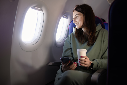 Portrait of a beautiful young woman in an airplane drinking coffee and using smartphone