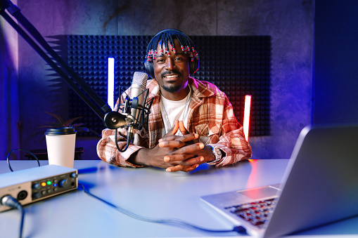 African radio host sitting at desk recording in studio with microphone and laptop close up