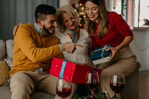 Shot of a happy couple bonding with a senior woman together and sharing wrapped gifts on the couch next to the Christmas tree.