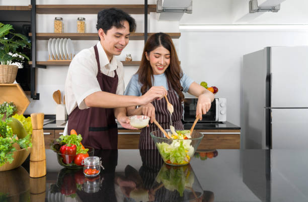 Young asian woman dressed in an apron mix the vegetables in a salad bowl together with a wooden ladle. Tomato, bell pepper and lettuce. Her boyfriend help putting salad dressing. Young asian woman dressed in an apron mix the vegetables in a salad bowl together with a wooden ladle. Tomato, bell pepper and lettuce. Her boyfriend help putting salad dressing. adults to help them lose weight stock pictures, royalty-free photos & images