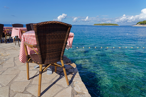 Set table of a restaurant on the Croatian Adriatic, on a stone wall, directly above the blue, crystal clear sea. In the background green overgrown islands and cloudy sky.