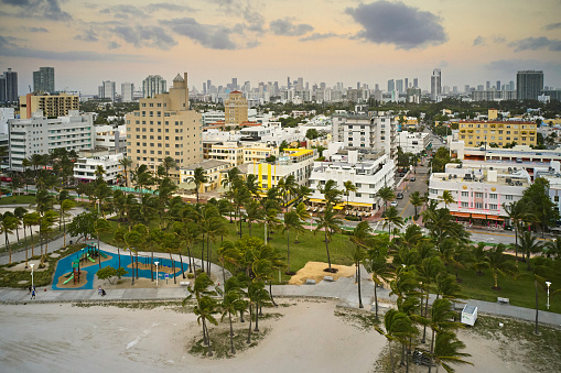 Drone point of view above beachfront with wind blowing in palm trees, Ocean Drive famous for art deco architecture, and downtown Miami in background.