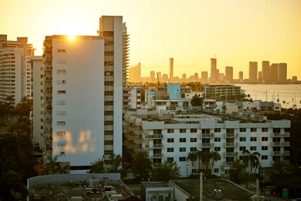 Drone point of view with focus on foreground residential buildings and hotels, bright sun, Biscayne Bay, and cityscape in background.