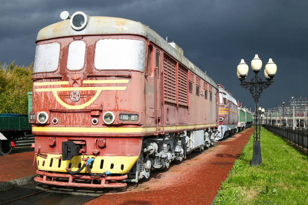 A green freight locomotive with white stripes is parked on a railway track, surrounded by greenery. The train's design and color scheme evoke a sense of industrial strength, while the natural backdrop contrasts with its mechanical structure.