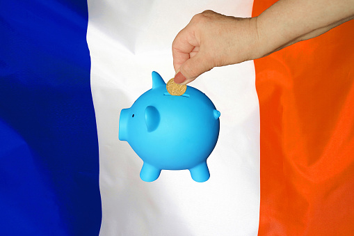 Hand of elderly woman putting coin into piggy bank on France flag background. Hand putting coin to piggy bank. Retirement savings, piggy bank. Concept saving money and retirement fund in France