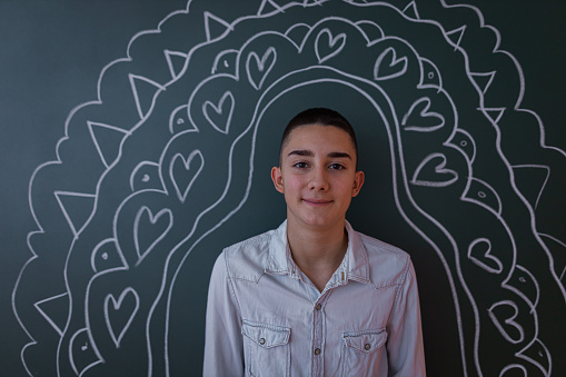 A portrait of a male young student wearing a white button shirt, standing by the chalkboard with chalk-drawn ornaments.