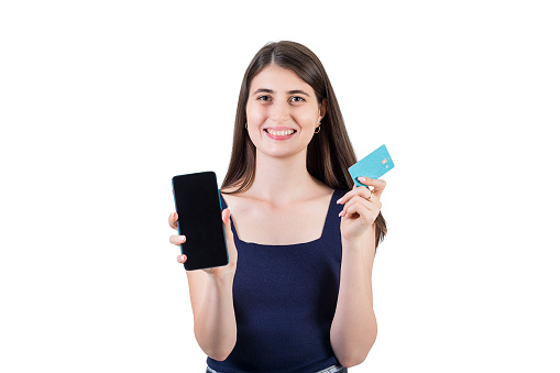 Positive young woman looking excited to camera showing her phone screen and holding a credit card in other hand, isolated on white background with copy space. Internet mobile banking concept