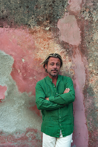 Blond tanned man in green shirt and white pants stands in front of weathered pink plaster wall.