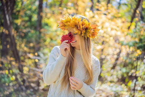 Outdoors lifestyle close up portrait of charming blonde young woman wearing a wreath of autumn leaves. Smiling, walking on the autumn park. Wearing stylish knitted pullover. Wreath of maple leaves.