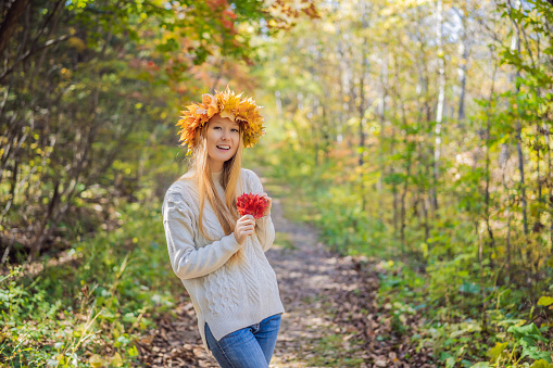 Outdoors lifestyle close up portrait of charming blonde young woman wearing a wreath of autumn leaves. Smiling, walking on the autumn park. Wearing stylish knitted pullover. Wreath of maple leaves.