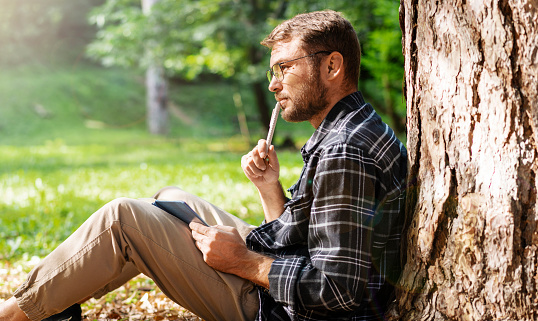 Adult man with eyeglasses sitting in the park and writing in his diary.