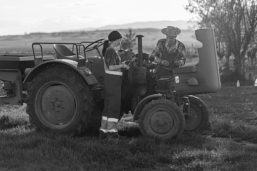 A black and white shot of an old tractor in the field, a daughter, and her farmer father fixing the vehicle.