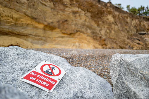 Shallow focus of a No Climbing sign attached to a shoreline boulder to help protect against coastal erosion. The distant cliffs are collapsing into the sea.