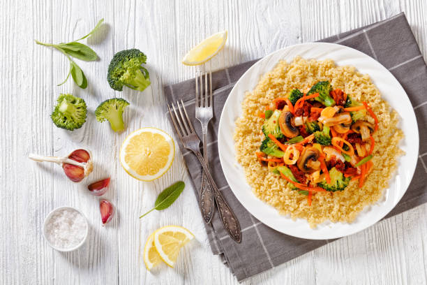 quinoa with stir fried broccoli, julienne carrots, sun dried tomatoes, leek and mushrooms on white plate on white textured wooden table with ingredients, horizontal view from above, flat lay