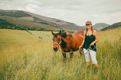 Amazon woman walking in the countryside with a brown horse. Cowgirl woman is wearing brown cap, black t-shirt, white pants and riding boots. Green landscape is seen with houses in the background in a rural area.