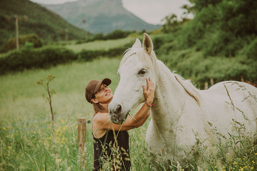 A woman in a meadow with tall grass petting a white horse.