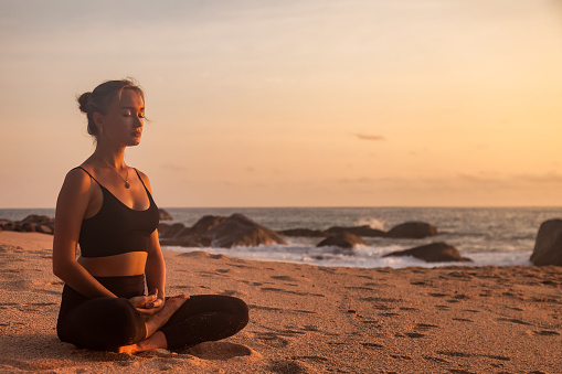 Young woman sitting does asana yoga position on tropical sea coast sandy beach, relaxing at sunset. Female performs exercises for healthy lifestyle to restore strength, spirit. Copy text space