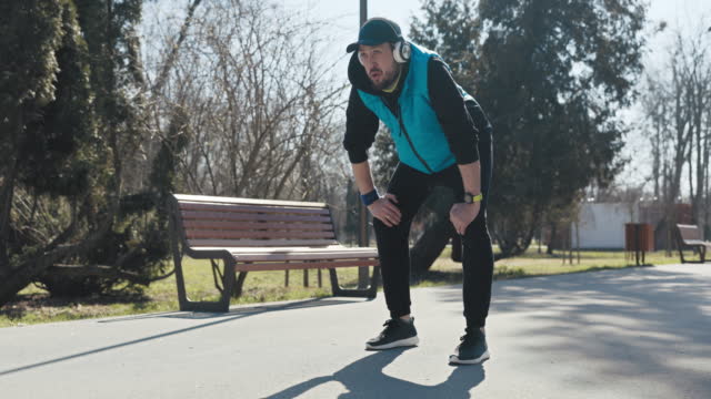 Young man runs in the park in the morning. He is dressed in black sports clothes, has cap on head and headphones. The athlete runs the last lap. He stopped, rested hands on knees and was breathing