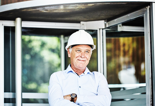 Proud middle-aged man with the white hard hat representing management stands outside a commercial building.