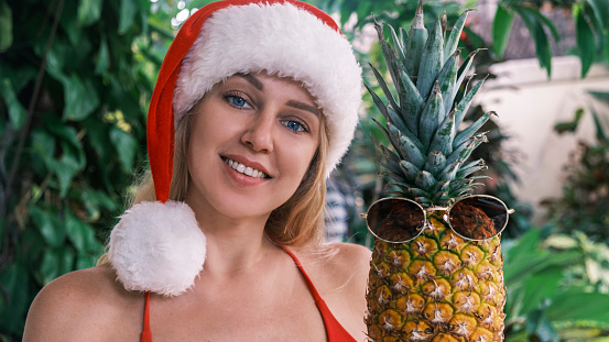 Beautiful smiling blonde woman in santa hat looking at camera holding pineapple wearing sunglasses in green tropical plants background. Concept of Christmas on tropical island. Festive card