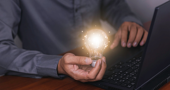 The hand of a man with a light bulb and he is using a notebook computer. Thinking differently creates inspiration and ideas.New innovations and technologies can always come from creative and different ideas.
