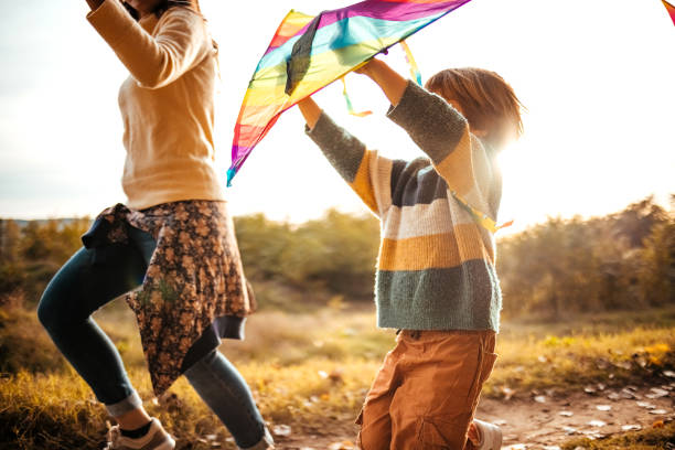 A helping hand. Caring mother teaches little girl to fly a kite in autumn nature at sunset. Child development, learning, freedom, joy, & connecting with nature.