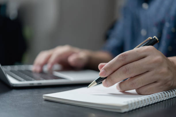 istock male hand writing in notebook with pen on laptop computer in modern office. 1431708061