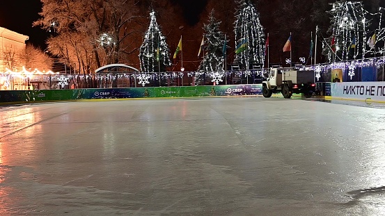 Ulyanovsk, Russia - January 6, 2022: An ice making resurfacer machine (filling machine) cruises by on the ice skating rink against the backdrop of Christmas decorations and twinkling garlands.