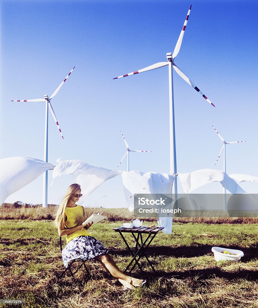 Smart Energy Smart young woman showing an alternative approach. She has her laundry dried in the wind, near wind power plant. In the meantime she enjoys her book and a tea. Wind Turbine Stock Photo