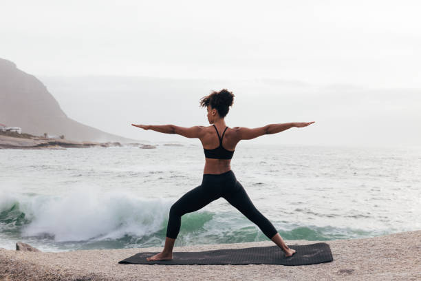 Back view of female practicing yoga in warrior pose by the ocean at bay stock photo