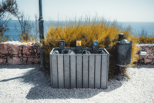 A wide-angle shot of three trash bins for separate waste collection: yellow for packed trash, blue for paper, and black for unidentified on the coastal pavement with a bush and the ocean behind it