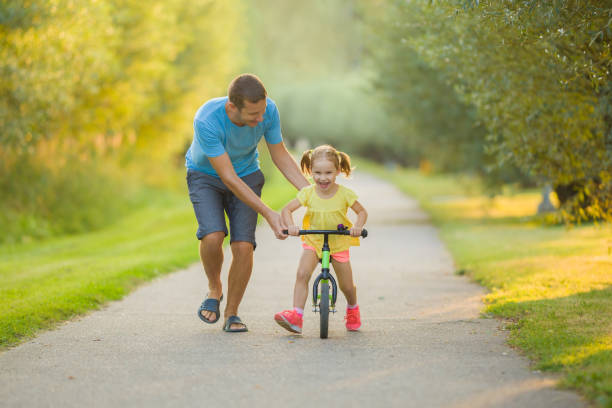 Young adult father teaching happy beautiful little girl to ride on first bike without pedals on sidewalk at city park. Learning to keep balance. Warm summer day. Cute 3 years old toddler. Front view. stock photo