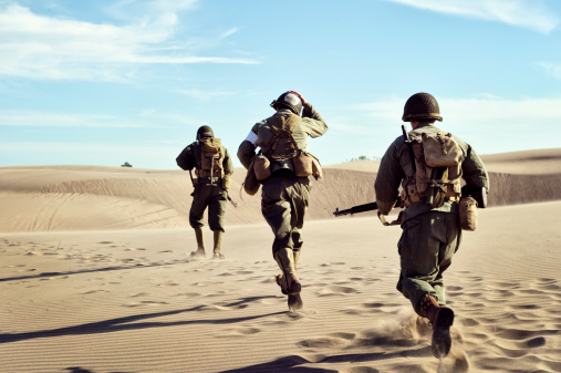 Three WWII Soldiers Running across the desert sand.  Uniforms, helmets, boots, gear, shovels, ammo belt, guns are all authentic for the time period.  Late afternoon shot.  Blowing desert sand in lower third of image creates a line across the picture.