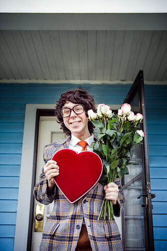 A nerdy young teenage man with a big red valentines day card / box of chocolates and a bouquet of roses stands excited, happy, and full of joy in front of his dates house, a goofy smile on his face.  Retro 1970's style clothes, hair, and glasses.  Wide angle shot in front of bright blue house.  Vertical with copy space.