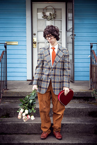 Sad Nerd with Valentine Gift and Flowers on Front Porch A nerdy young man with a big red valentines day card / box of chocolates and a bouquet of roses stands by the front door of a bright blue house after just being rejected by a potential date.  Retro 1970's style clothes, hair.  Bright blue house; Vertical. face down stock pictures, royalty-free photos & images