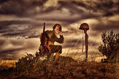 Soldier in full combat uniform kneeling At the marked, temporary grave of another soldier.  Gun stuck in ground with bayonet.  Helmet gun, bayonet and strap clearly visible in ominous sky.