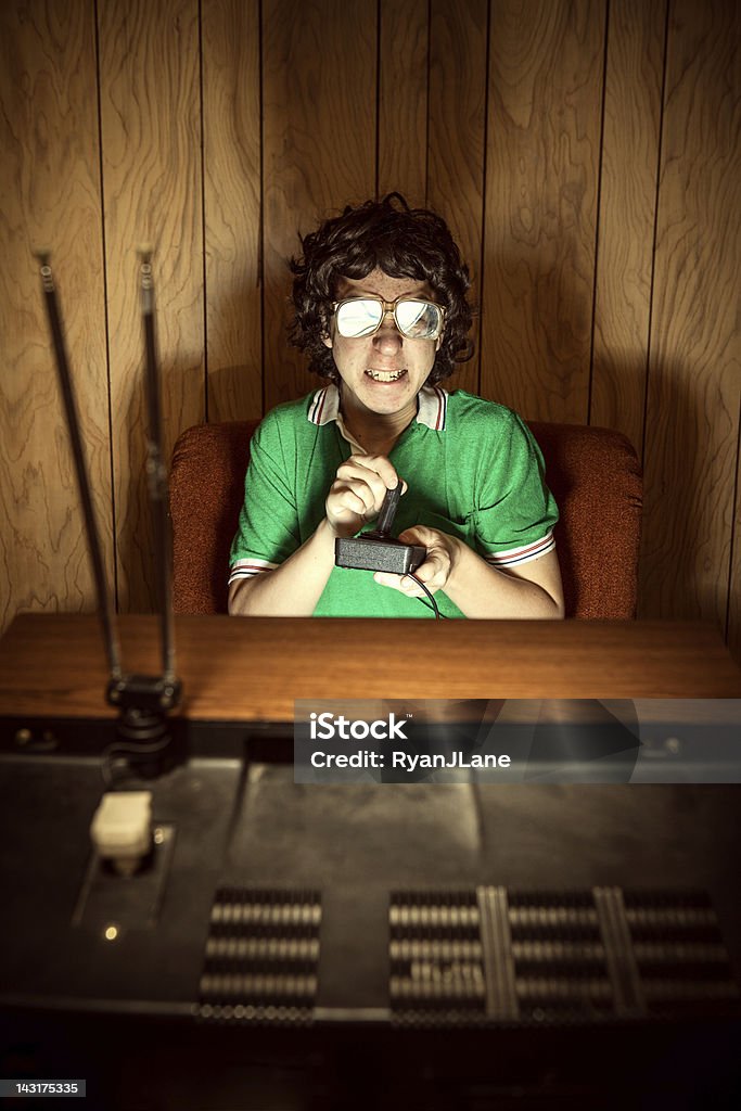 Gamer Nerd Playing Video Games on T.V. A curly haired retro styled 1980's teenager plays a video console games on his television, gritting his teeth in concentration as he spaces out in his living room.  Vintage wood paneling on the walls.  Vertical with copy space. Video Game Stock Photo