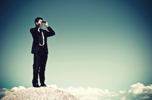 Businessman holding binoculars on boulder over the clouds.