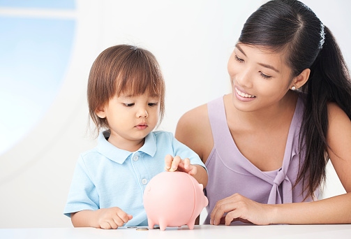 Cute boy putting a coin into his piggy bank with mother by his side. 