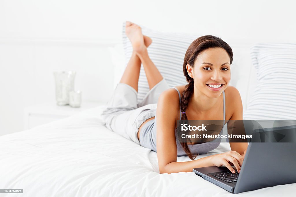 Giovane donna sdraiata sul letto utilizzando un computer portatile - Foto stock royalty-free di Abbigliamento casual