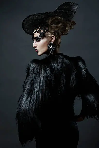 Side view of beautiful blond young woman fashion model, posing in a black fur coat and elaborate hat with updo. Avant Garde makeup.