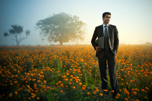 A confident Indian businessman with laptop standing in marigold farm field.