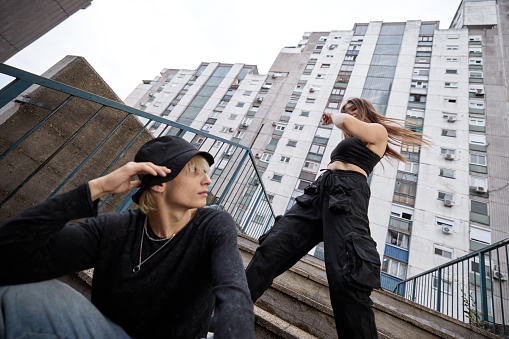 A teenage girl is dancing surrounded by the buildings while a teenage boy is looking at her.