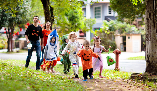 Child in Halloween costume. Mixed race Asian and Caucasian kids and parents trick or treat on street. Little boy and girl with pumpkin lantern and candy bucket. Baby in witch hat. Autumn holiday fun.